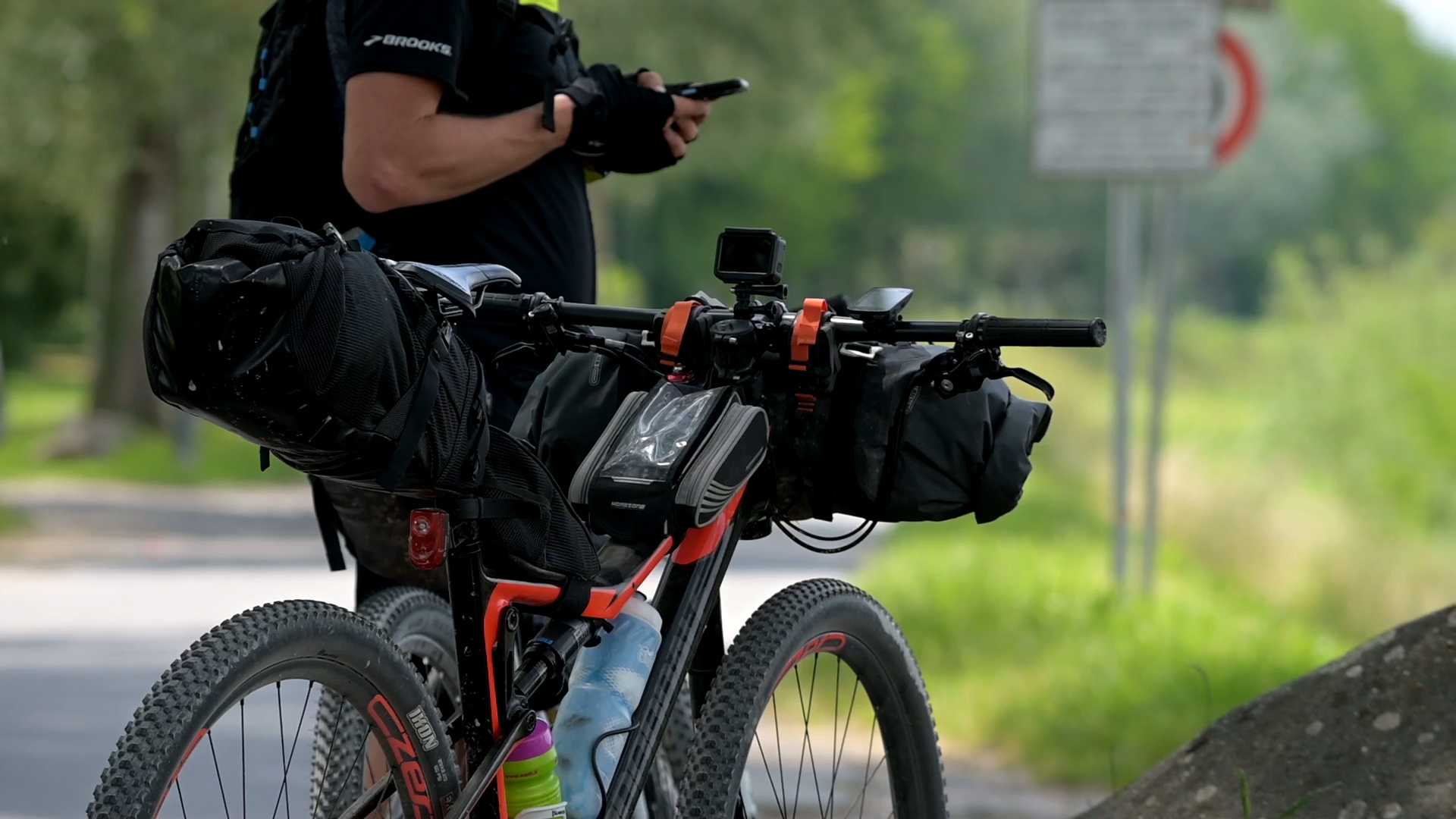 Primo piano di una bicicletta caricata con borse per cicloturismo, ferma lungo una strada. Il ciclista è in piedi vicino alla bicicletta e sta consultando il cellulare.