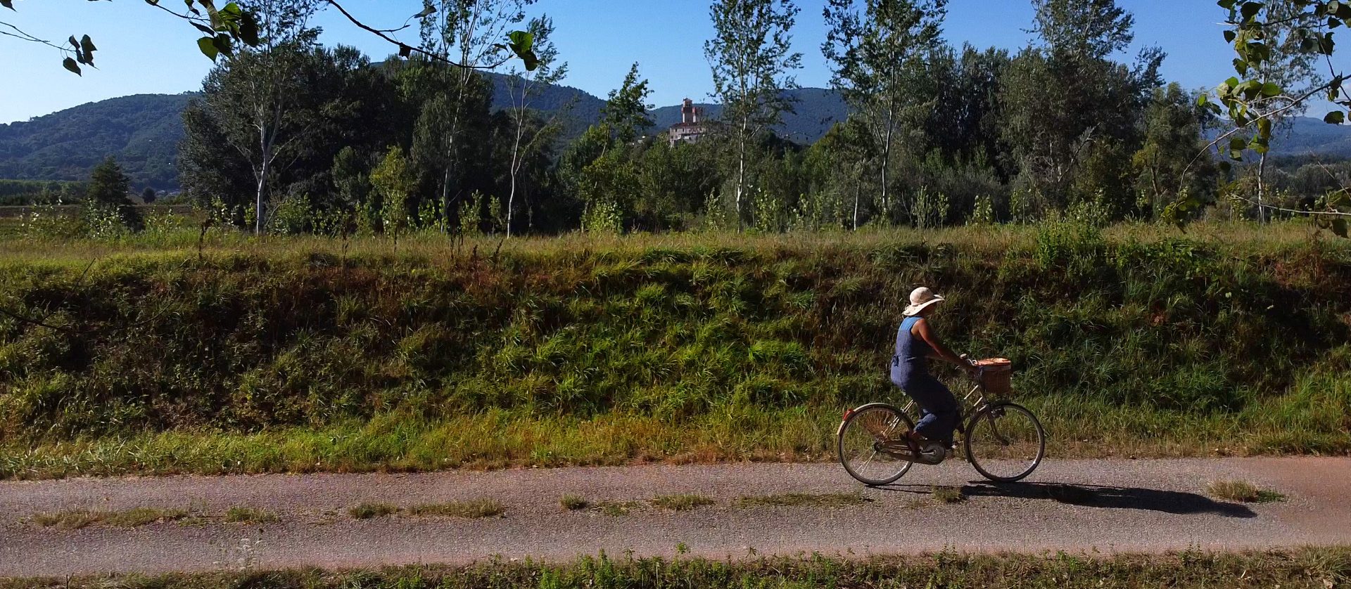 Una donna con un vestito azzurro e un cappello bianco in bicicletta sugli argini del Serchio. Oltre gli argini ci sono dei pioppi e il castello di Nozzano.