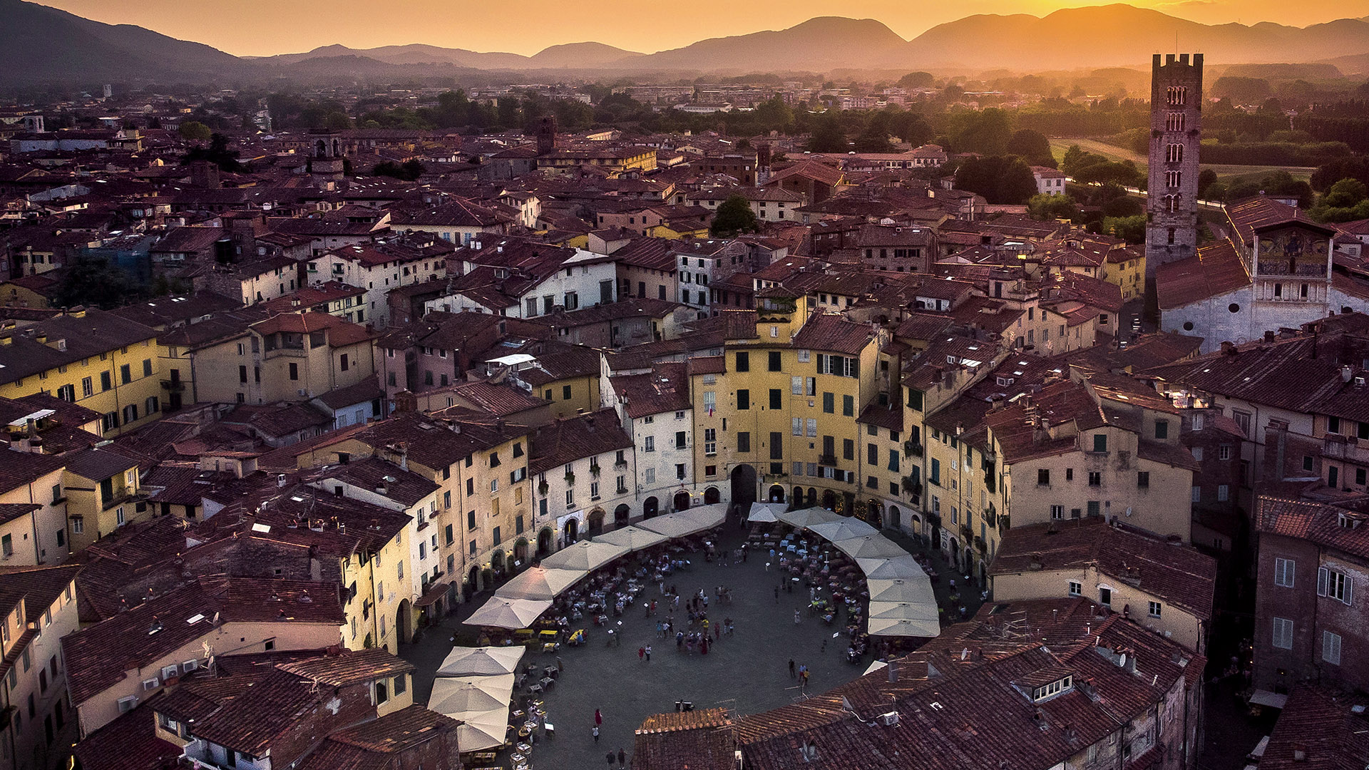 Panorama dall'alto di Piazza dell'anfiteatro a Lucca, tra i tetti delle case, nella luce del tramonto. Sullo sfondo le colline.