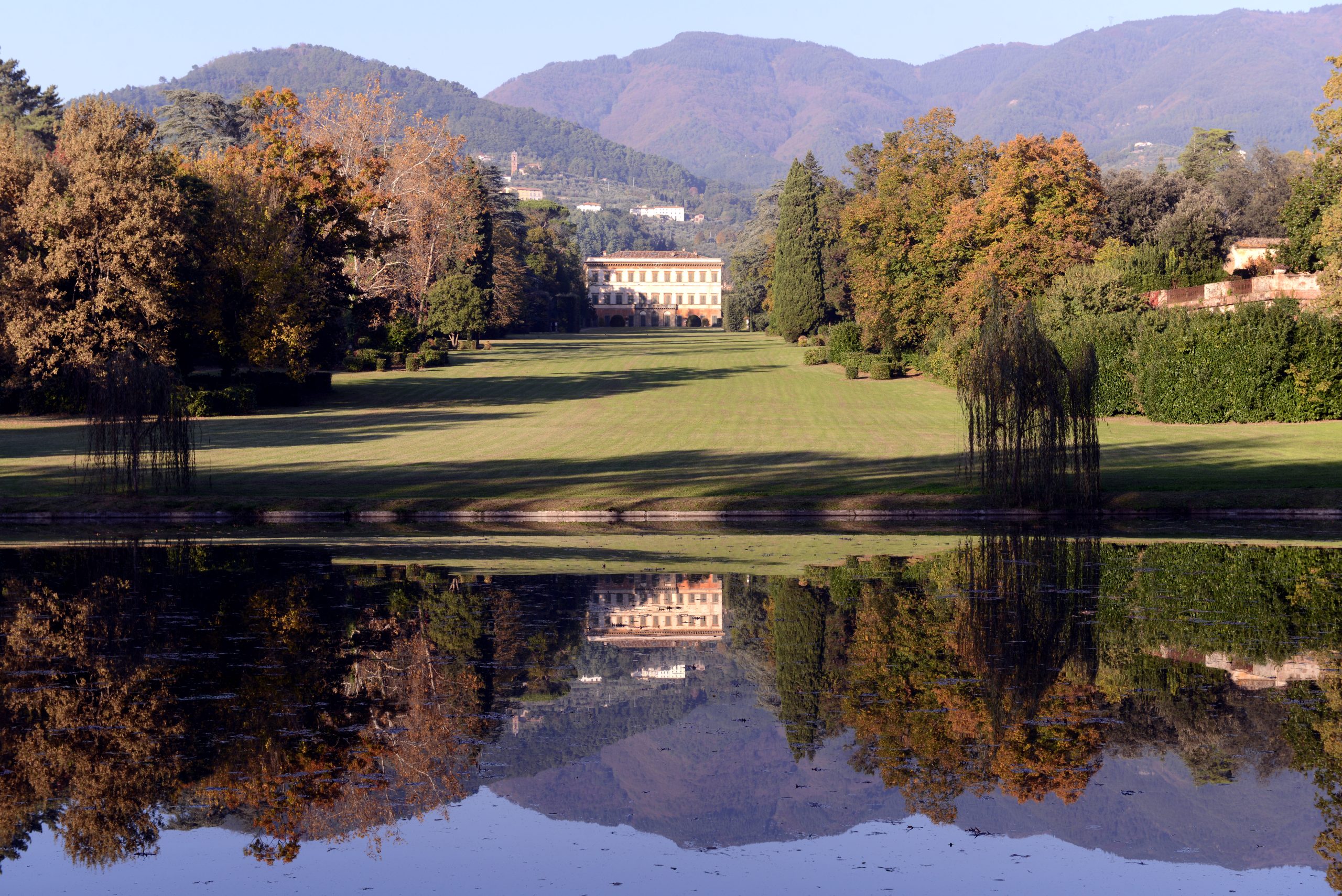 Panorama di villa Reale con la villa che si rispecchia nel laghetto. Ai lati della villa ci sono alberi in abito autunnale e sullo sfondo montagne