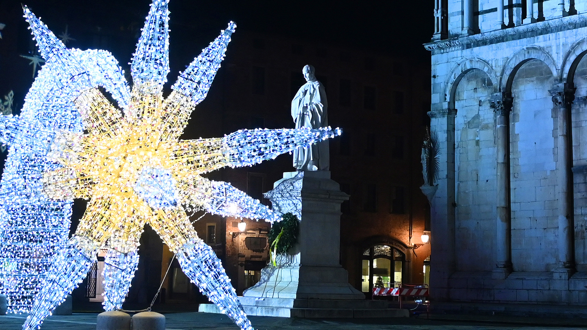 In primo piano una grande stella fatta di luci in piazza San Michele, per Natale. In secondo piano la statua di Francesco Burlamacchi e dietro la chiesa di San Michele