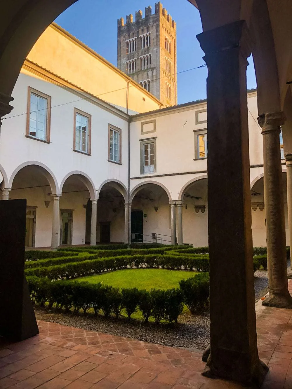Angolo del chiostro del Real collegio di Lucca con il campanile della basilica di San Frediano. Al centro del chiostro il giardino con aiuole.
