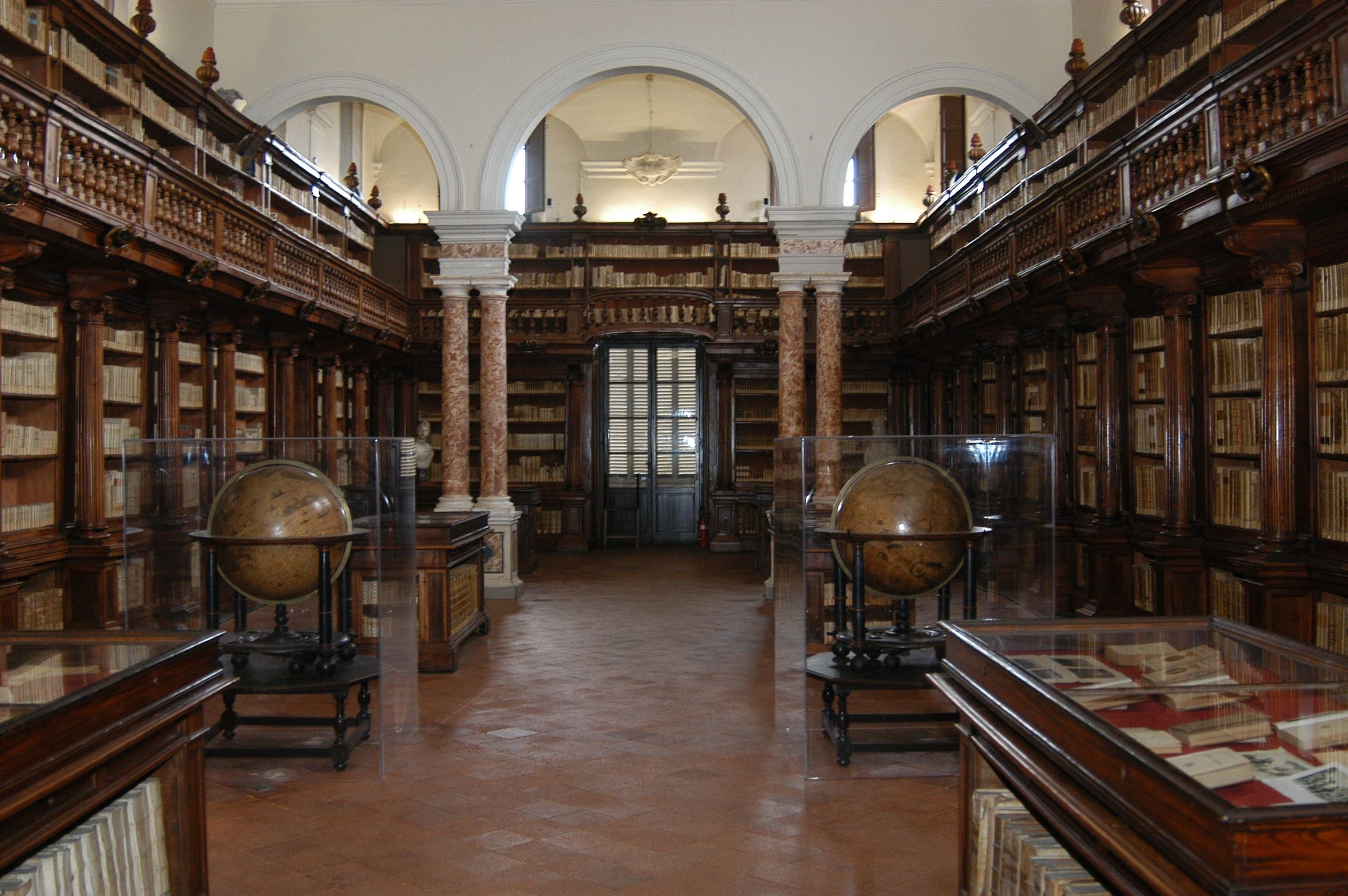 prospettiva della sala dei mappamondi della biblioteca statale di Lucca. Sul fondo una arco a serliana e ai lati scaffali con libri
