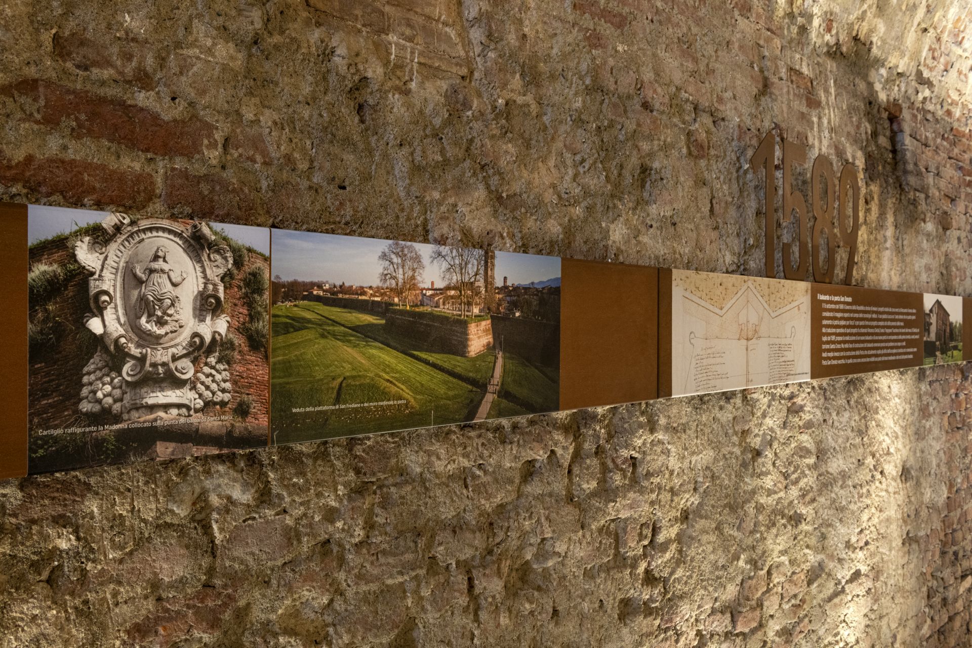 parete del sotterraneo del baluardo santa croce con una fascia metallica suilal quale sono sitemate foto e didascalie sulla storia delle mura di lucca