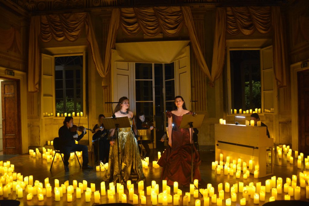 due donne in abito da sera stanno in piedi davanti a un quartetto d'archi nel salone centrale di villa bottini illuminato da candele poste sul pavimetno.