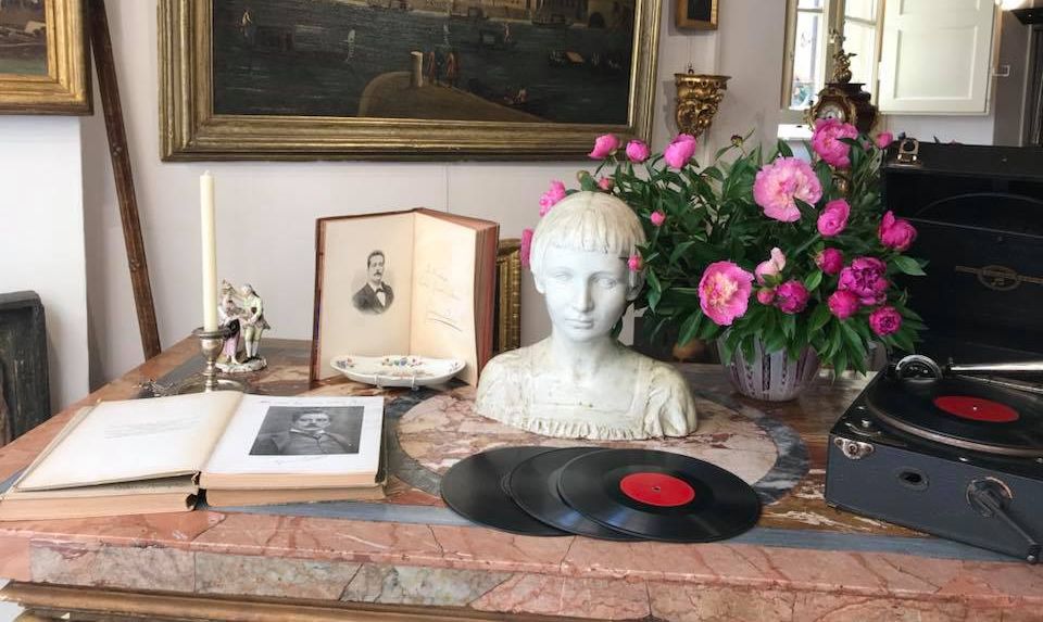 vbetrina di un negozio di antiquariato. Su un tavolo di marmo ci sono un libro aperto, alcuni dischi in vinile il busto di una ragazza, un vaso con fiori