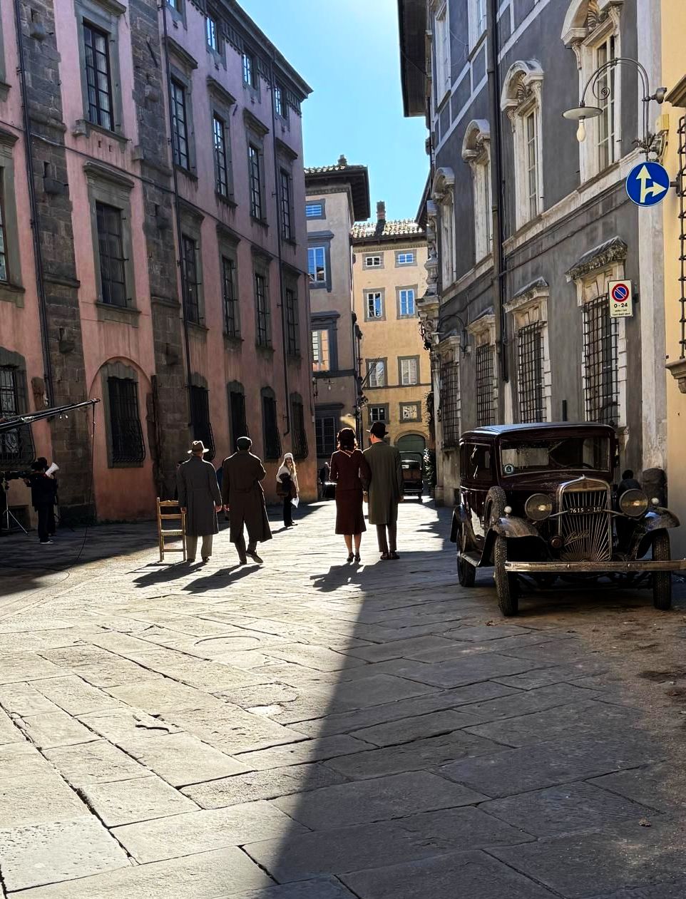 due grandi palazzi settecenteschi si affacciano sulla strada in cui passeggiano due attori in abiti degli anni '50. sulla destra è parcheggiata un auto d'epoca