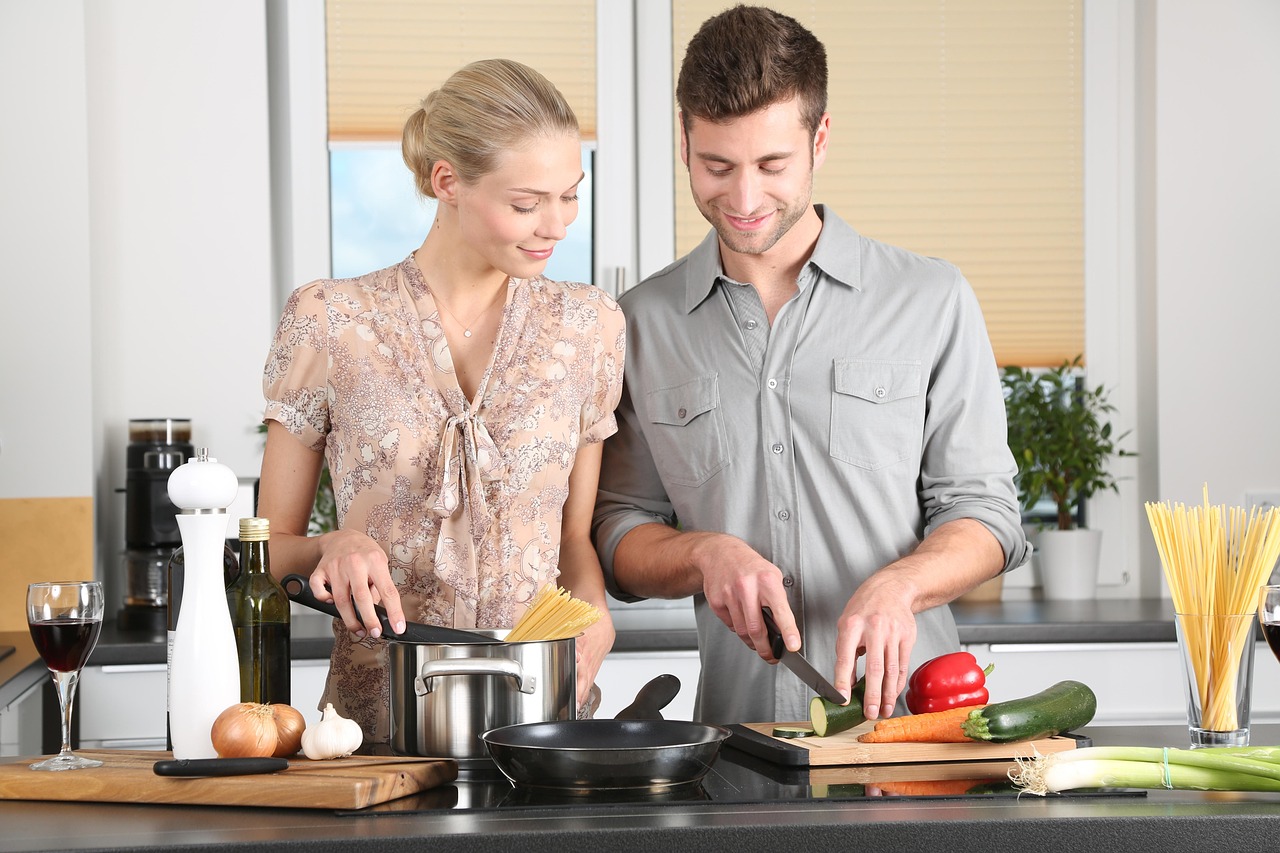 un ragazzo e una ragazza davanti al tavolo di una cucina preparano da mangiare. Sul tavolo ci sono attrezzi da cucina, verdure e un bicchiere di vino rosso. Lui affetta verdure, lei lo guarda e prepara una pentola con spaghetti.