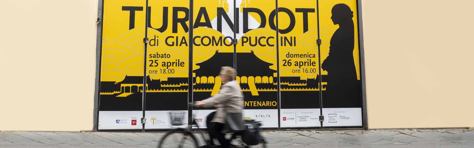 una signora in bicicletta passa davanti all'uscita degli artisti del teatro del giglio dove campeggia un grande manifesto con sritto turandot 100 l'anniversario
