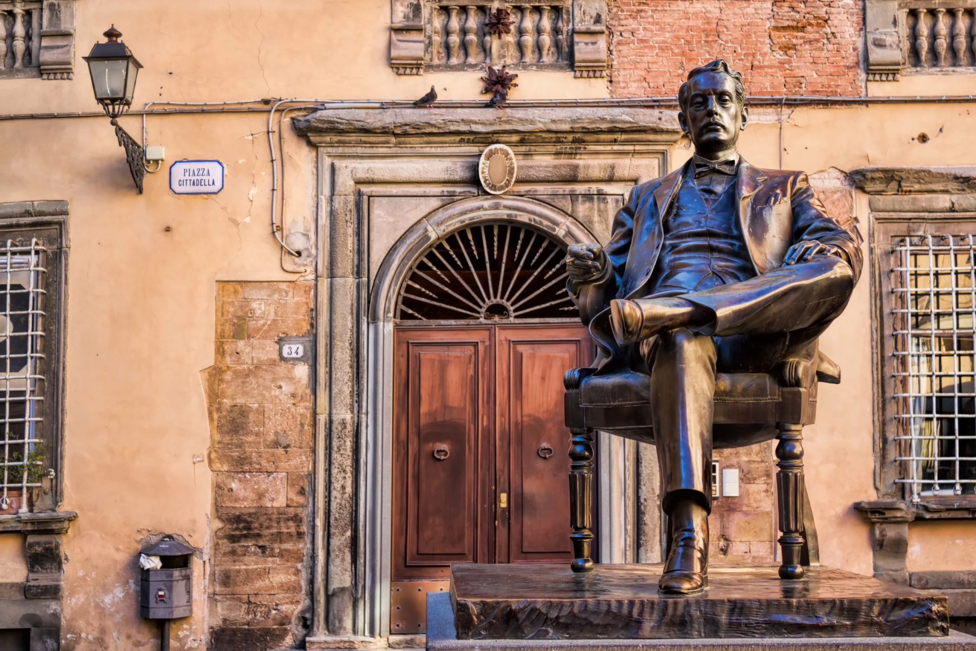 Statua di bronzo Giacomo Puccini in piazza Cittadella. Puccini è seduto su una poltroncina con una gamba accavallata e un sigaro in mano. Sullo sfondo palazzi storici.
